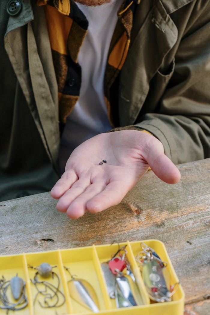 A person holds fishing hooks in hand beside a tackle box on a wooden surface.